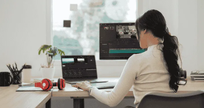 Person editing a cooking video on a laptop, with headphones and a second monitor in a bright workspace.