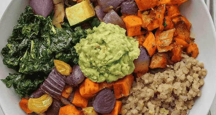 Close-up of a colorful roasted veggie grain bowl with sweet potatoes, kale, quinoa, and avocado mash.